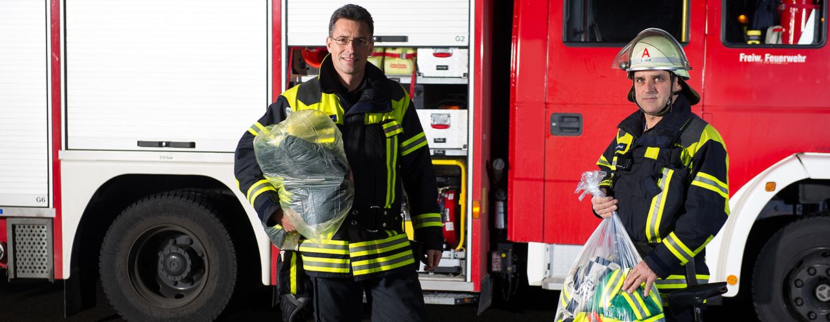 Zwei Feuerwehrleute vor einem Löschfahrzeug mit verpackter Einsatzkleidung in wasserlöslichen Wäschebeuteln